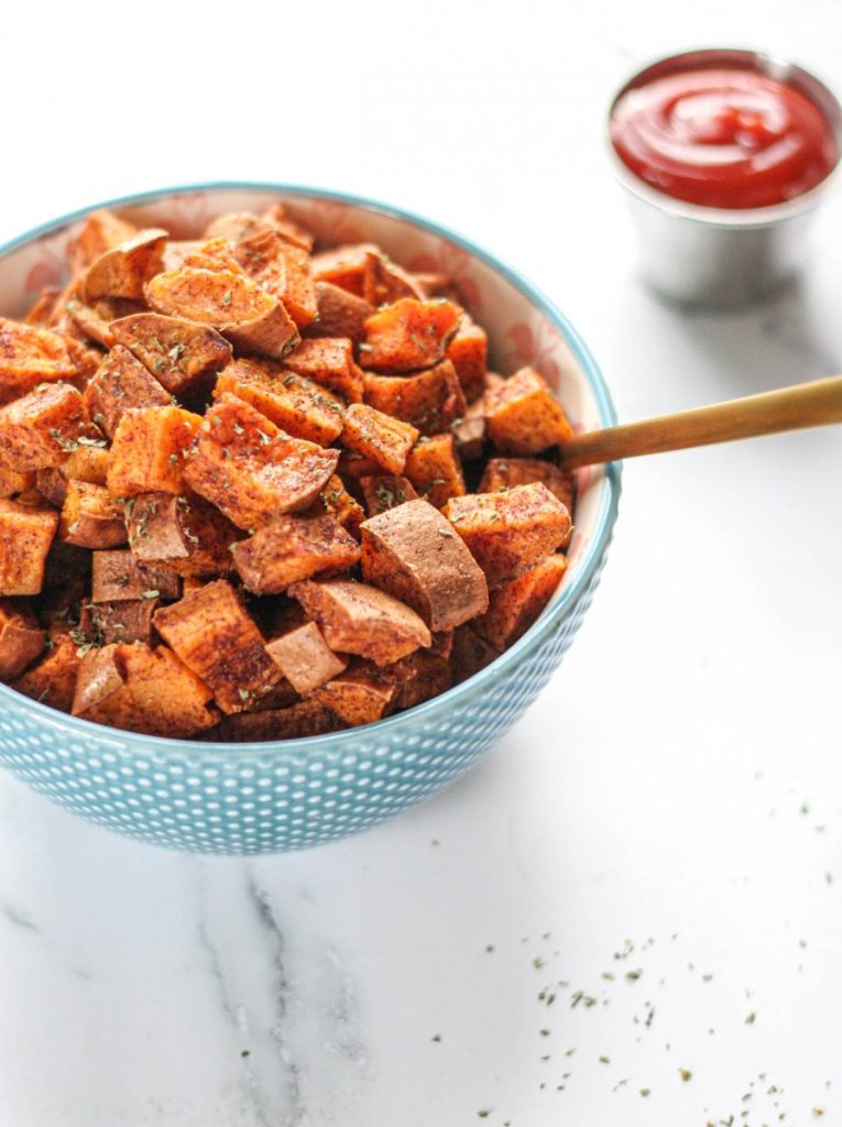 Blue bowl of Roasted Cinnamon Sweet Potatoes with a gold serving spoon and a ramekin of ketchup on a white counter top