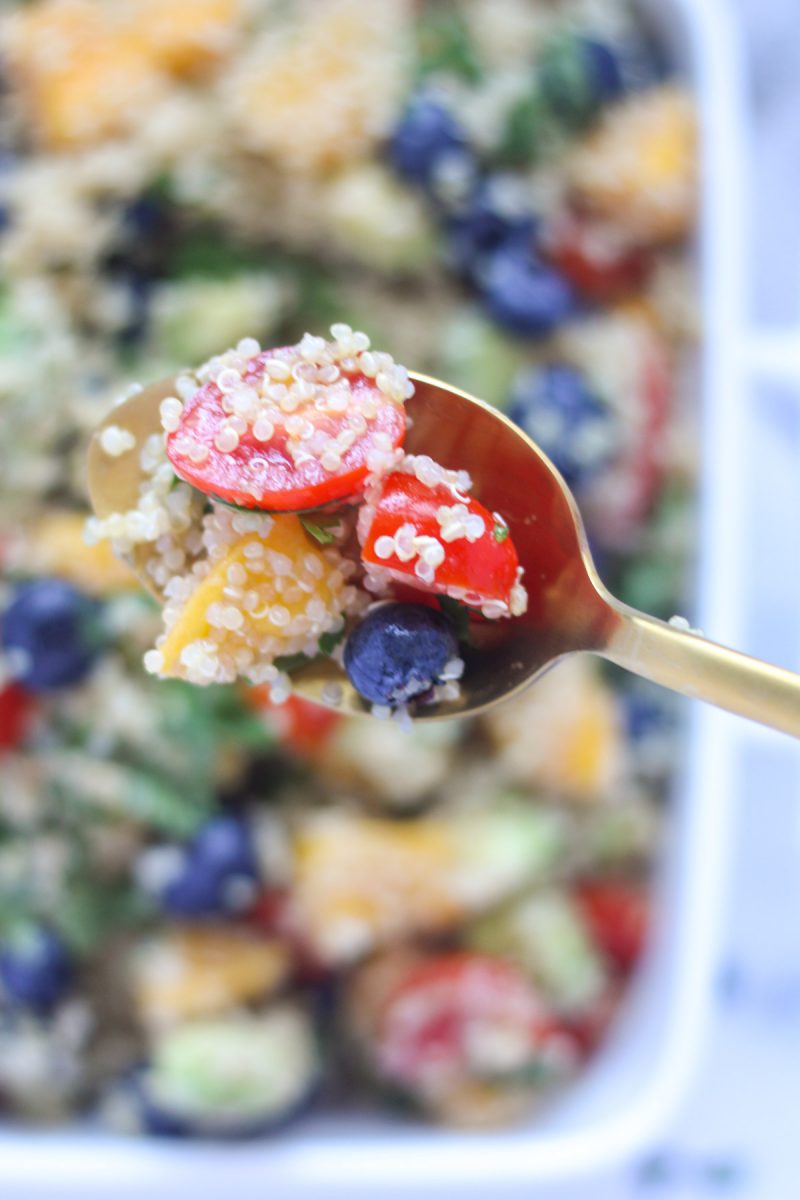 A gold spoon with cherry tomatoes, a blueberry and a piece of mango on it, covered in some quinoa Summer Quinoa Salad