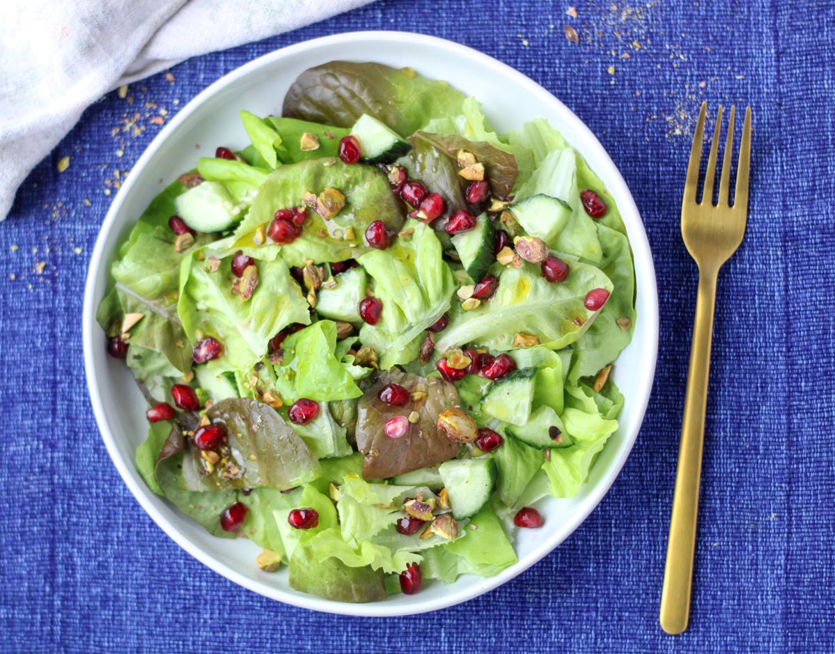 A large white plate filled with Butter Lettuce and Pomegranate Salad on a blue tablecloth with a gold fork beside