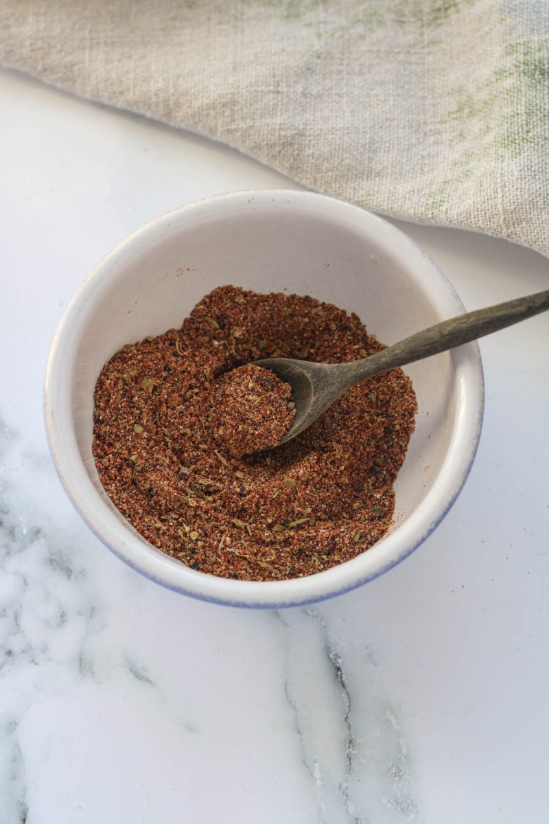 A small bowl of taco seasoning with a brown spoon on a white countertop