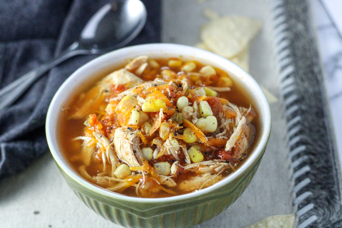 A small green bowl of chicken soup with corn and shredded carrots with a silver spoon on the side