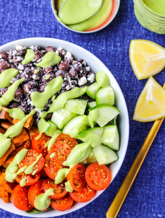 A white bowl filled with black beans, sweet potatoes and veggies on a blue tablecloth and lemon wedges beside