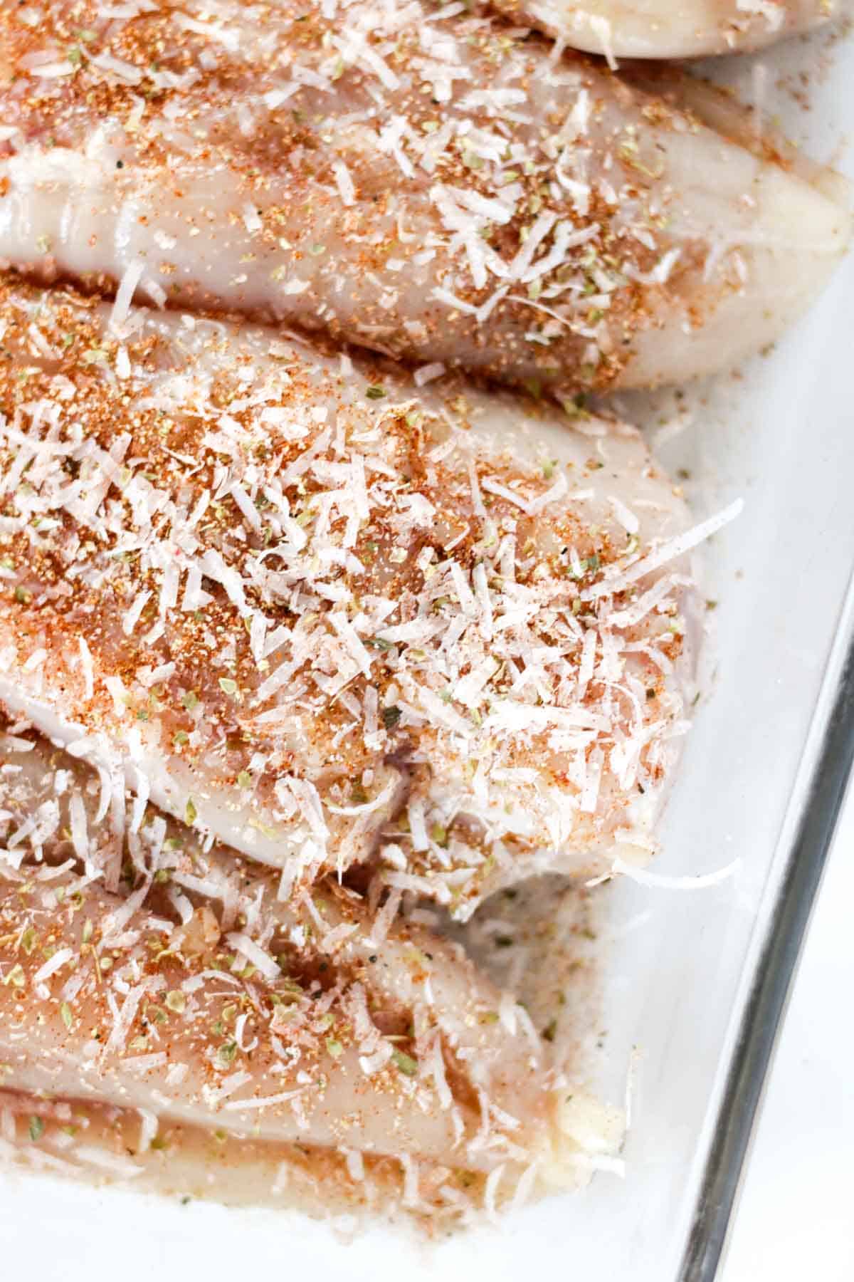 Chicken breasts covered with parmesan cheese and herbs in a glass baking dish.