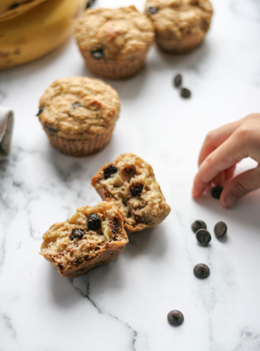 Best Ever Banana Chocolate Chip Muffins on a white countertop with a child's hand stealing a chocolate chip