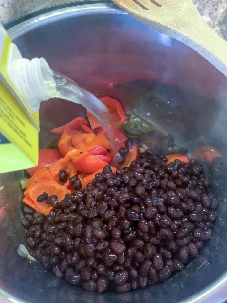 Chicken broth being poured into Instant Pot filled with black beans and roasted red peppers