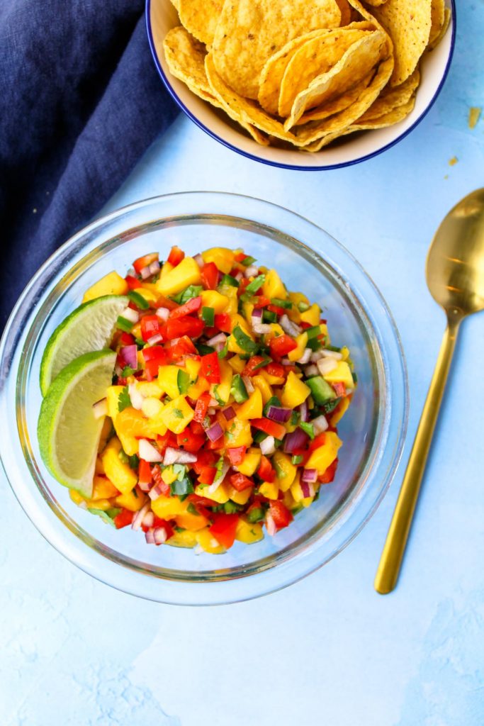 A glass bowl filled with mango salsa with a gold spoon beside.