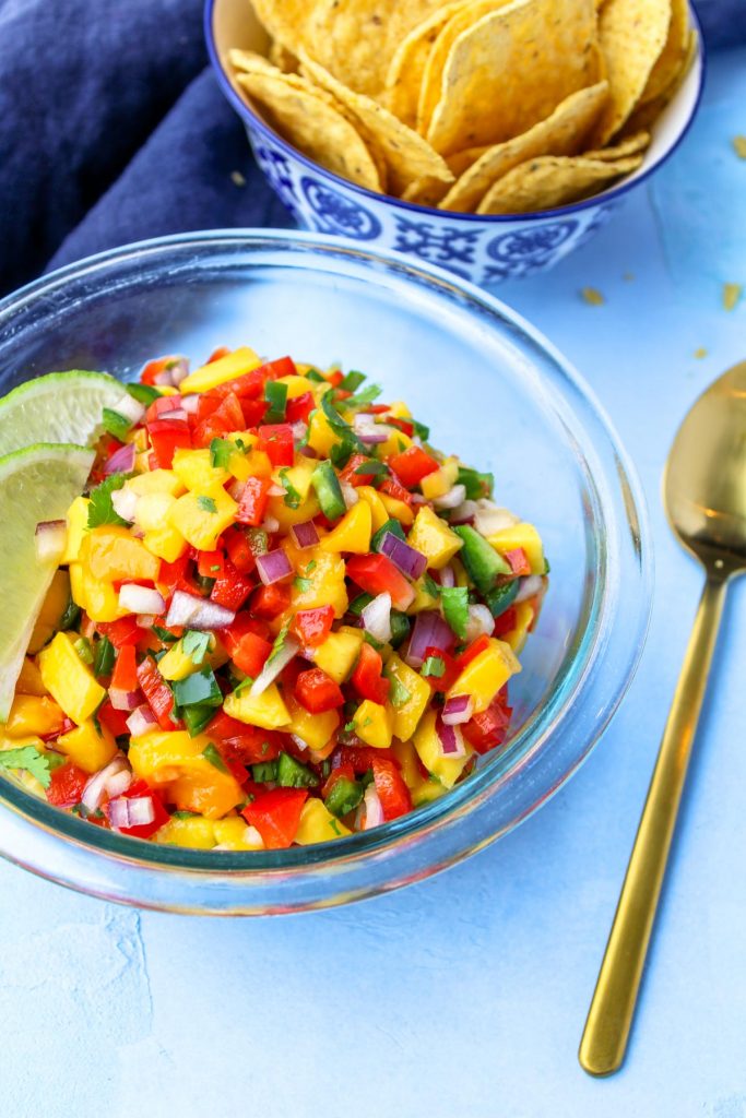 A glass bowl filled with mango salsa with a gold spoon beside.