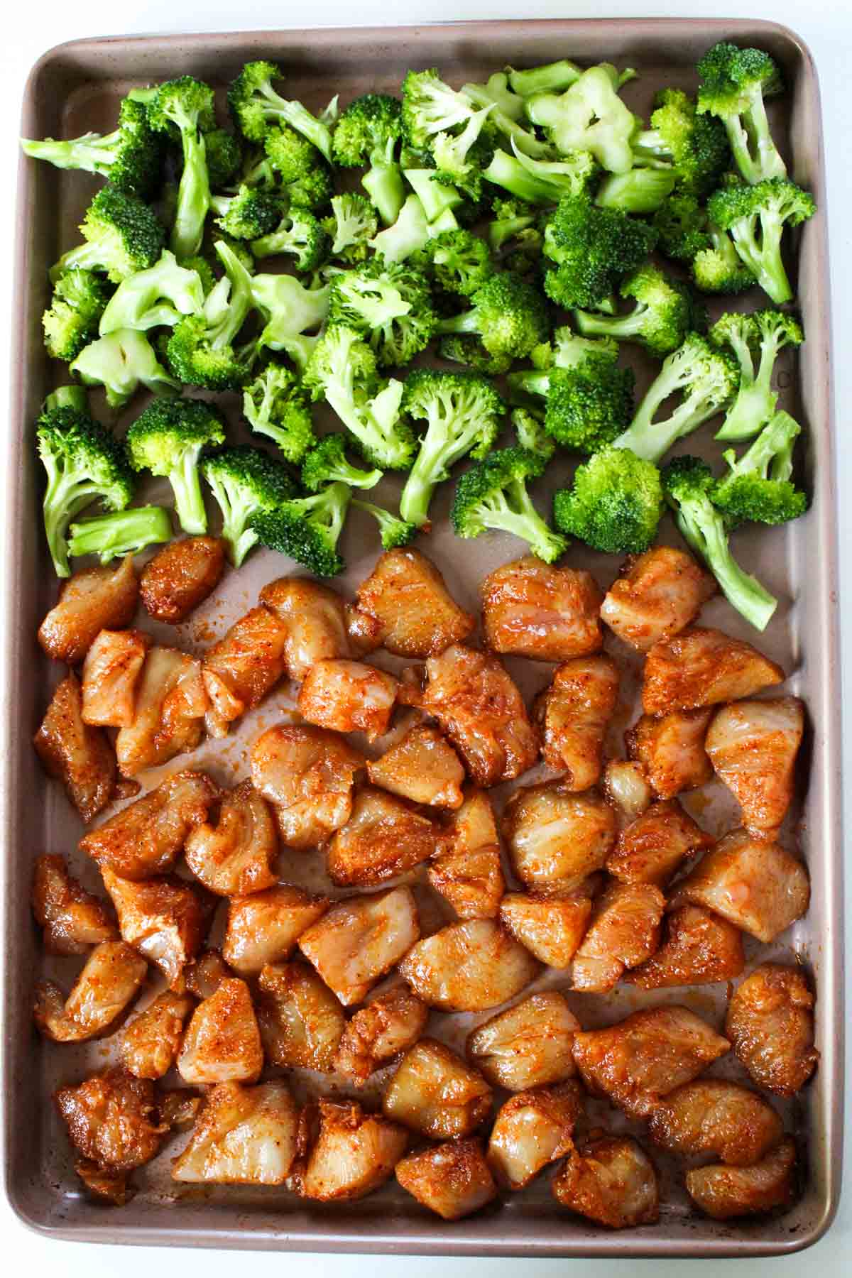 Large sheet pan covered with marinated chicken breast chunks and broccoli florets.