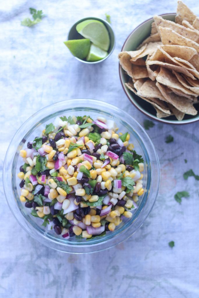 Summer Corn and Black Bean Salsa in a clear glass bowl with a bowl of chips and lime wedges on the side
