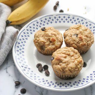 Three Best Ever Banana Chocolate Chip Muffins on a white plate with chocolate chips and bananas in the background