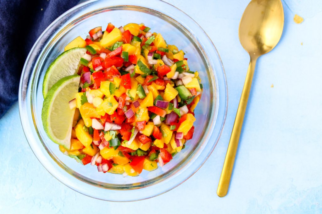 A glass bowl of chopped mango salsa with a gold spoon beside.